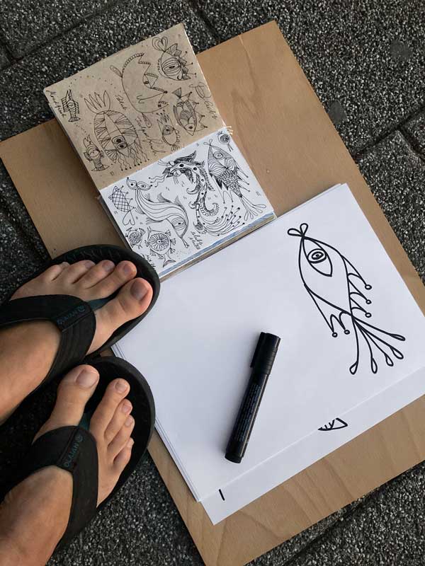 Tabea Heinicker hat ihre Zeichnungen auf den Boden gelegt. Links ihr kleines Reiseskizzenbuch mit verschiedenen Fischzeichnungen und rechts eine Reinzeichnung mit Edding auf Papier.