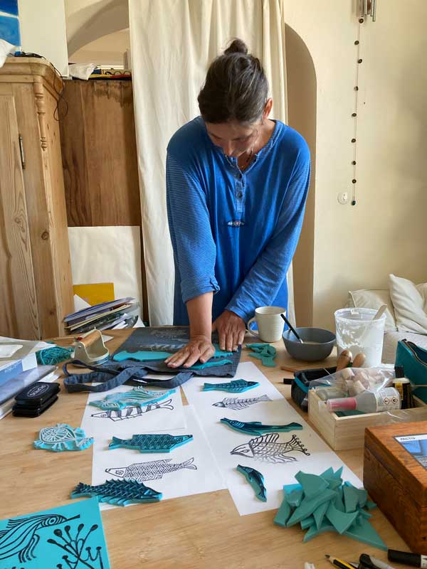 Therese Olivier beim Drucken von Fisch verliebten Stempeln auf Jutebeutel. Der Tisch ist voller Druck- und Arbeitsmaterial.