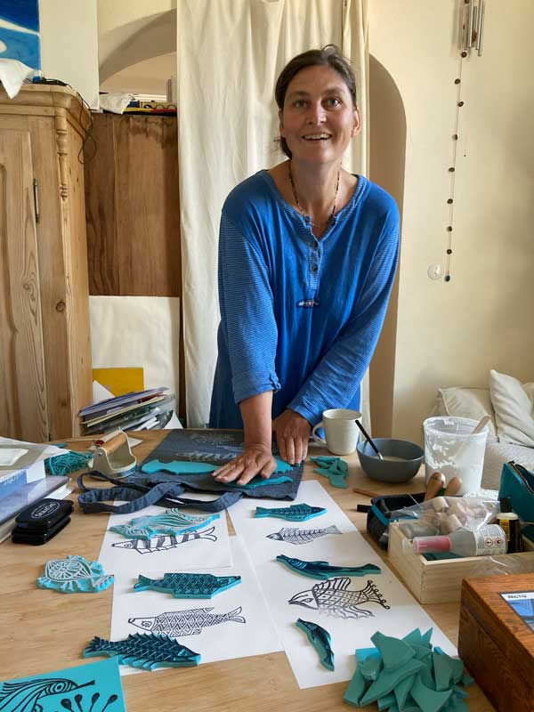 Therese Olivier beim Drucken von Fisch verliebten Stempeln auf Jutebeutel. Der Tisch ist voller Druck- und Arbeitsmaterial.