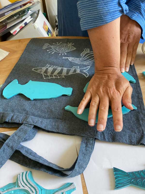 Nahaufnahme von Therese Oliviers Hand beim Drucken. Verschiedene Fisch-Stempel werden mit Weiß auf einen grauen Beutel gedruckt.
