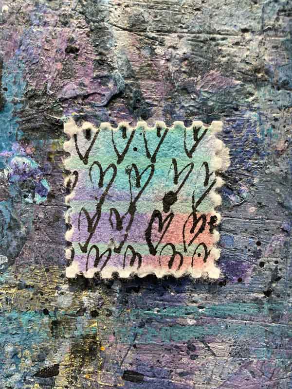 Eine Briefmarke auf der Arbeitsplatte mit vielen kleinen Tuscheherzen auf buntem Büttenpapier. Türkis, Lila, Koralle und Salbeigrün.