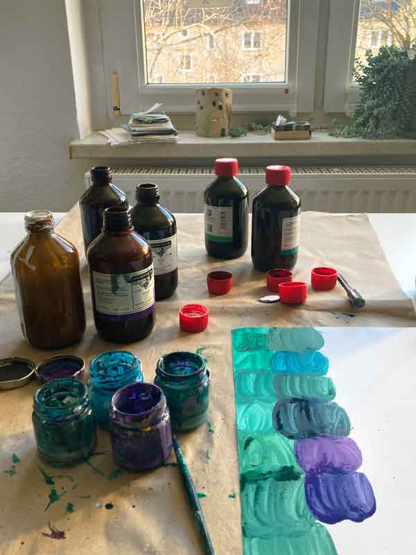 Tuscheflaschen und angemischte Farbgläschen mit Farbproben in Grün und Lila auf dem Schreibtisch.