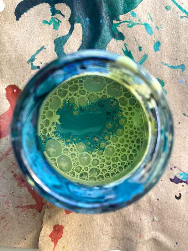 Blick in einer Tuscheglas, in dem gerade ein neues Grün gemischt wird. Eine Mittelgrüntürkis-Pfütze schwimmt im Hellgrünen Schaum.