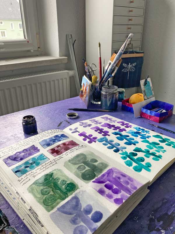 Foto vom Arbeitstisch mit aufgeschlagenem Skizzenbuch. Lila, Türkis und Graugrün. Im Hintergrund stehen Werkzeuge, Pinsel, Wasserglas, Tusche und Feder.