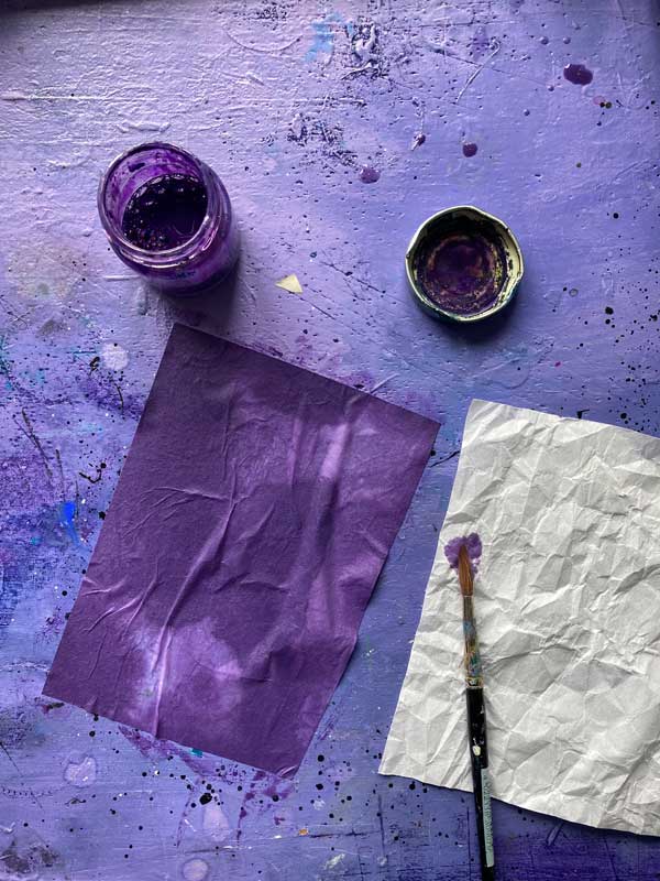 Tatulpen für die Frühlingspost 2025: Papierexperimente. Zu sehen ist ein zerknautschtes Linoldruckpapier und ein bereits lila eingefärbtes Papier auf dem lila Schreibtisch. Tuschefass und Pinsel stehen herum.