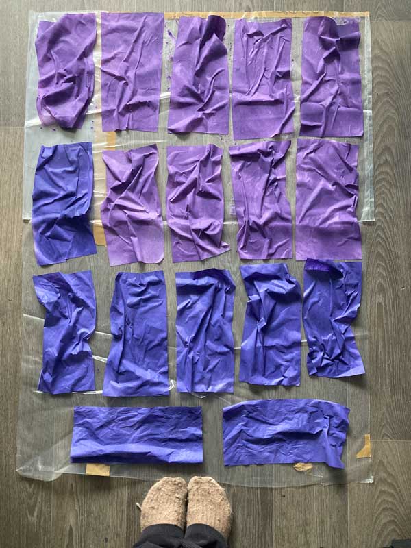 Blick auf den Fußboden aus Tabeas Perspektive. Ihre Füße sind zu sehen. Sie trägt hautfarbene Socken von Therese. Auf einer großen Folie liegen 20 eingefärbte Blätter. Die Blätter sind sehr schmalformatig und entweder Lila oder Blaulila. Die Papiere sind noch nass und extra zerknautscht.