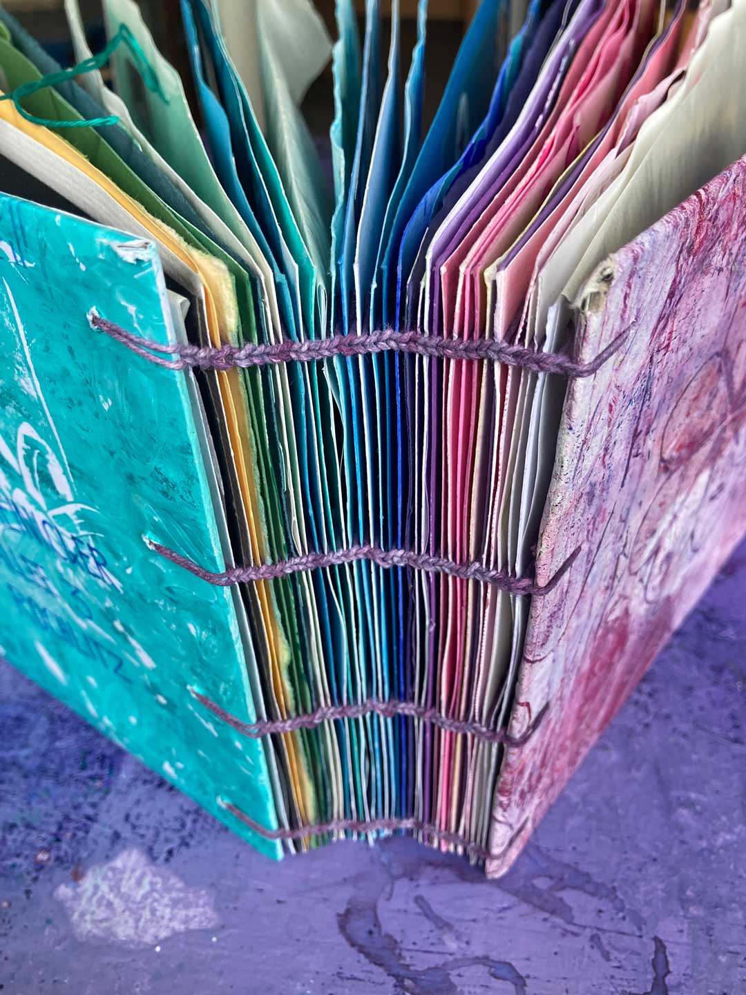 Ansicht des gebunedenen Buches aus allen Einsendungen der Frühlingspost 2025. Waisenbindung. Bunt im Regenbogen. 4 lila Kettstichzöpfe sind zu sehen.