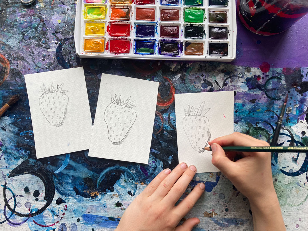 Von den Kindern lernen: Auf der bunten Tischplatte liegen drei Aquarellpapierchen mit gezeichneten Erdbeeren darauf. Kinderhände zeichnen an der dritten Erdbeere.
