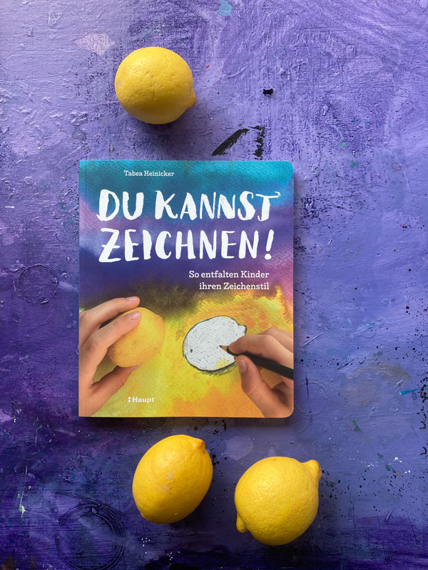 Das Buch »Du kannst zeichnen!« liegt auf einer lilafarbenen Schreibtischplatte. Drei Zitronen liegen um das Buch verteilt.