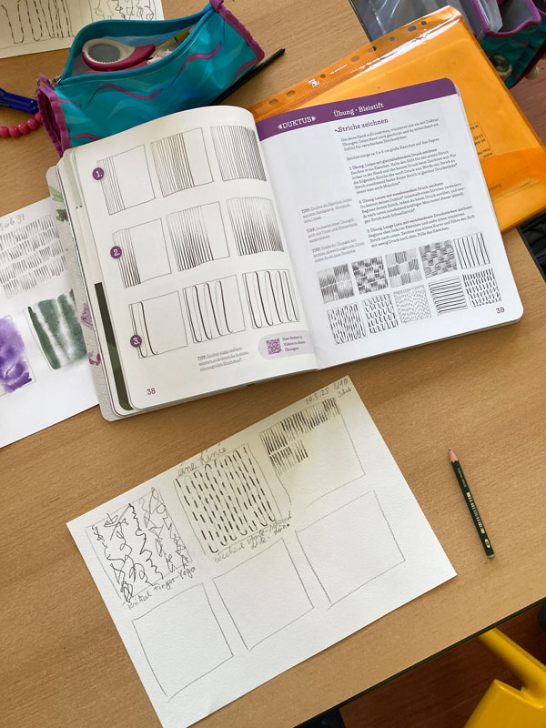 Das Buch liegt aufgeschlagen auf dem Schultisch. Duktus-Übungen sind zu sehen. Tabea hat den Kindern die Übungen vorgezeichnet, damit sie wissen, in welchem Tempo sie sie zeichnen.