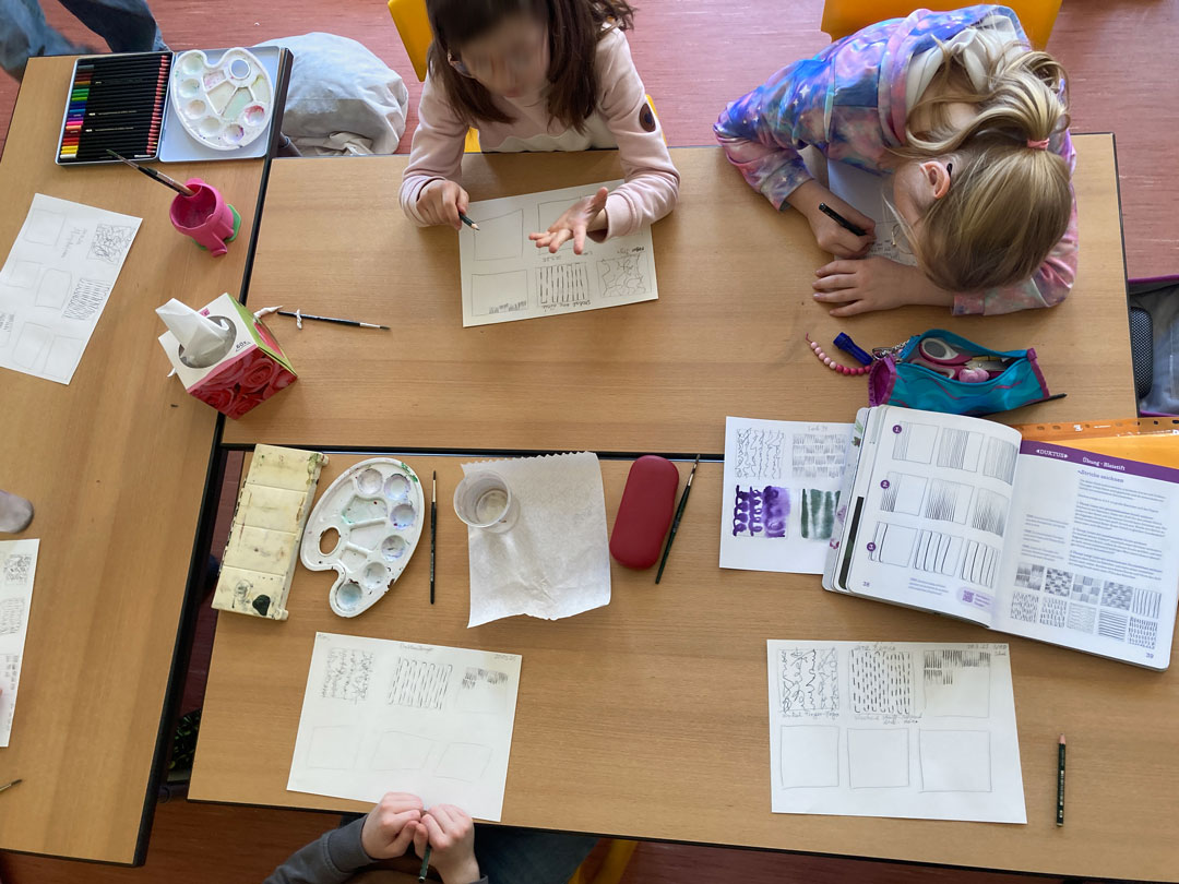 Von den Kindern lernen: Die Kinder sitzen am Tisch und zeichnen vertieft Duktus-Übungen. Tischaufsicht mit Buch auf dem Tisch.