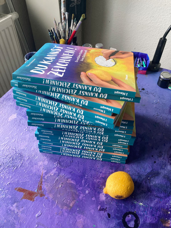 Ein bunter Stapel frisch gedruckter und ausgepackter Bücher »Du kannst zeichnen!« liegen auf Tabeas lilafarbenen Schreibtisch. Mit Zitrone!