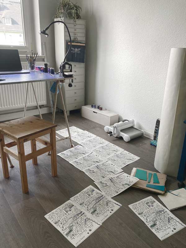 Foto von Tabeas Arbeitstisch. Die Bildbriefe liegen zum Trocknen auf dem Boden. Auch die vier Teile der Druckplatte liegen zum trocknen auf dem Fußboden.