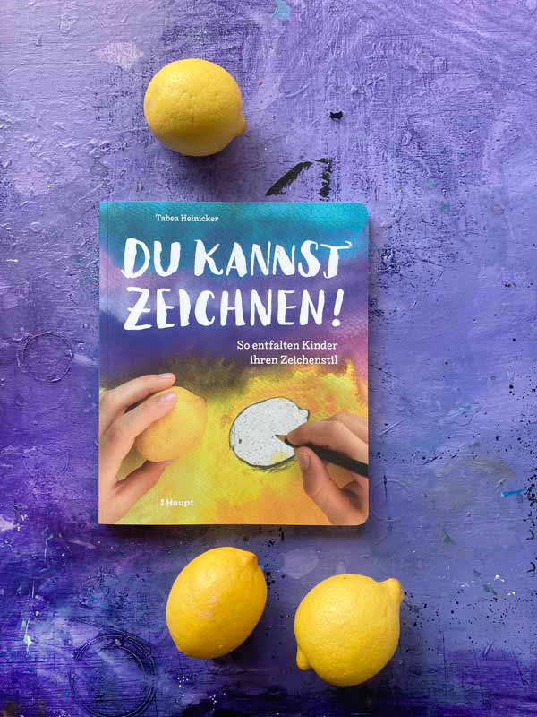 Das Buch „Du kannst zeichnen!« von Tabea Heinicker liegt auf Tabeas lilafarbener Arbeitsplatte. Drei frische Zitronen liegen um das Buch verteilt und ergänzen die Zitrone auf dem Buchtitel.
