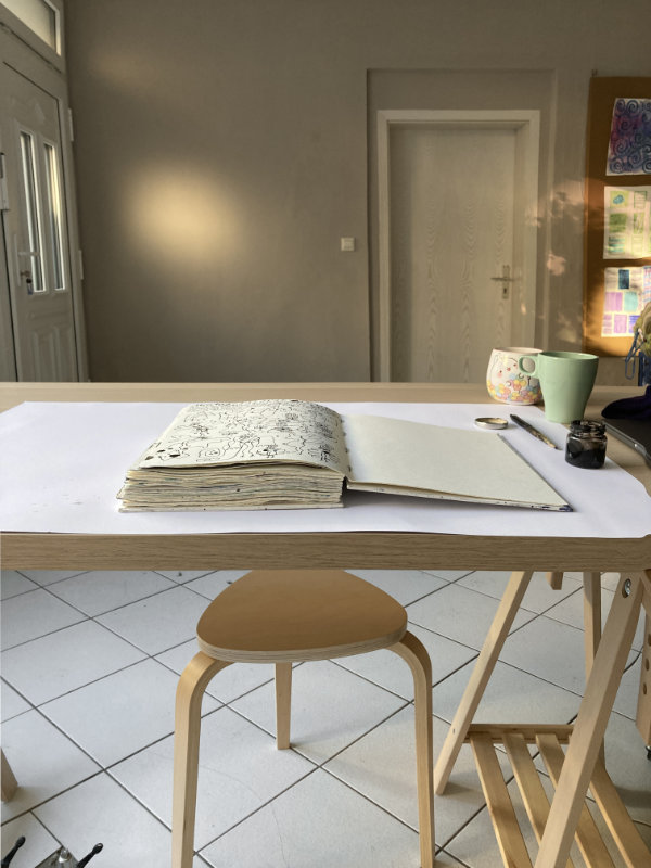 Tabeas Ateliertisch. Hocker und Tischplatte und Skizzenbuch. An der Wand Lichtspiele der Sonne.