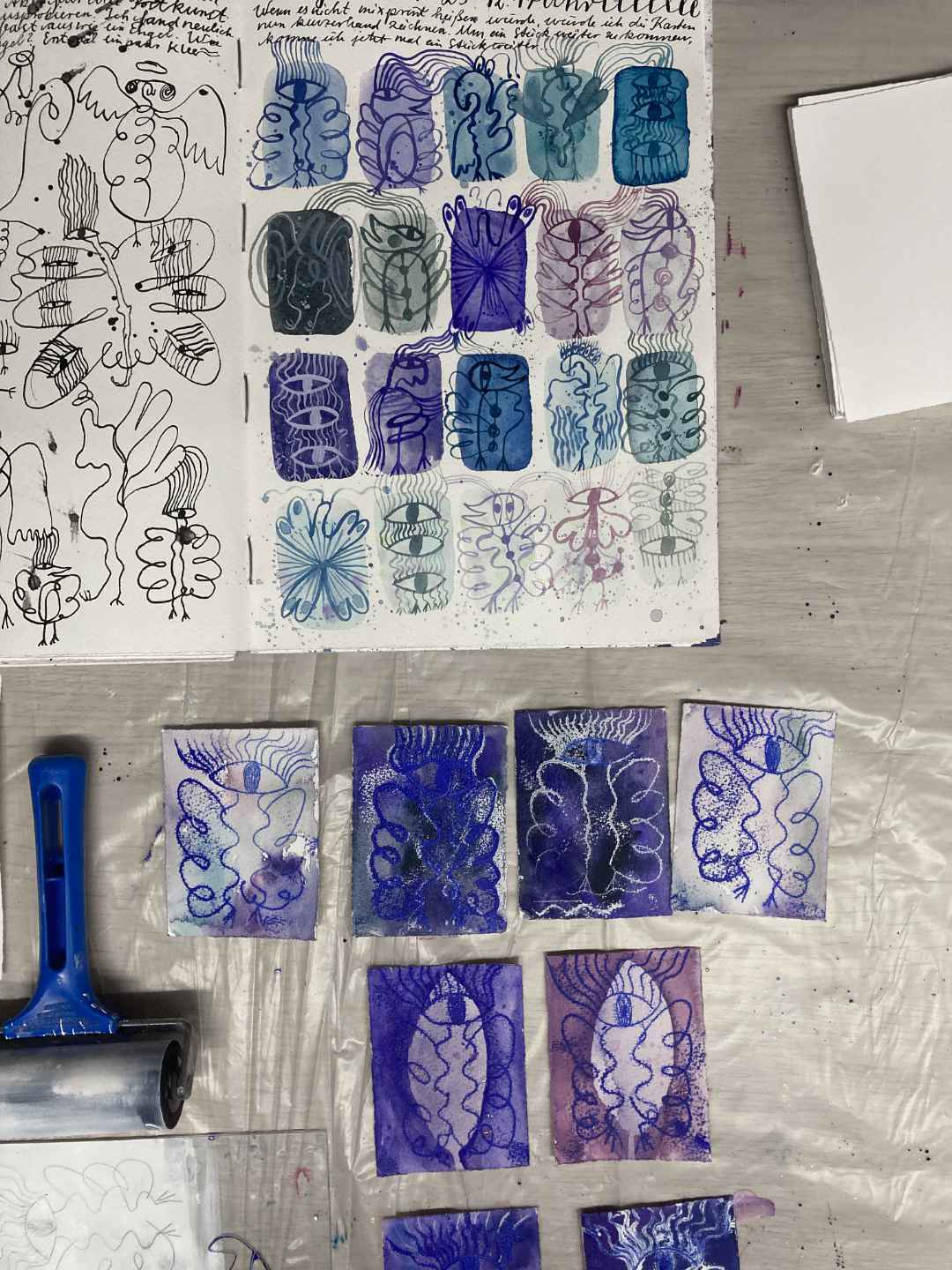 Engel im Mixed-Print: Arbeitstisch von obenfotografiert. Oben das Skizzenbuch mit den vielen kleinen Engeln in Farbe. Darunter erste Probedrucke in der Glasscheibendruck Technik. Farbwalze, Glasscheibe im Anschnitt.