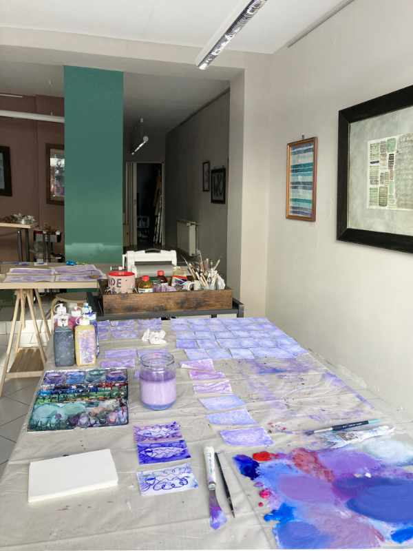 Blick in den Raum, mit Tabeas Arbeitstischen. Tisch ist voller Papierchen, die schon grundiert sind. Vorn liegt die Glasscheibe mit vielen Farbflächen, die angemischt wurden.