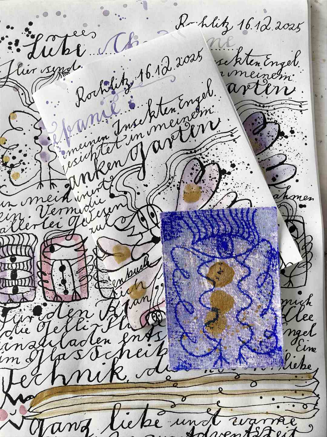 Aufsicht Bildbrief und Engeldruck. Im Hintergrund liegt ein kompetter Bidbrief, der koloriert wurde. Darauf liegt ein gefalteter Bildbrief und darauf ein blauer Engelsdruck.